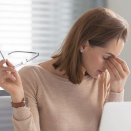 Fotolia_288393810: Frau vor einem Laptop mit einer Brille in der einen Hand fasst sich an die Stirn.