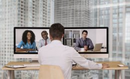 Mann im Business-Look sitzt vor zwei Bildschirmen und nimmt an einer Videokonferenz mit drei weiteren Personen teil
