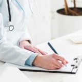Eine Ärztin sitzt an einem Schreibtisch und notiert etwas auf einem Klemmbrett