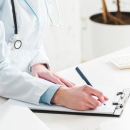 Eine Ärztin sitzt an einem Schreibtisch und notiert etwas auf einem Klemmbrett