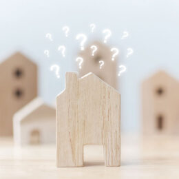 Ein Miniaturhaus aus Holz steht neben weiteren Holzhäusern auf einem Tisch. Über dem Haus sind Fragezeichen zu sehen.