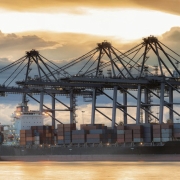 Schiffe werden von Kränen mit Containern beladen. Die Szenerie spielt sich im Hafen bei Abendrot ab